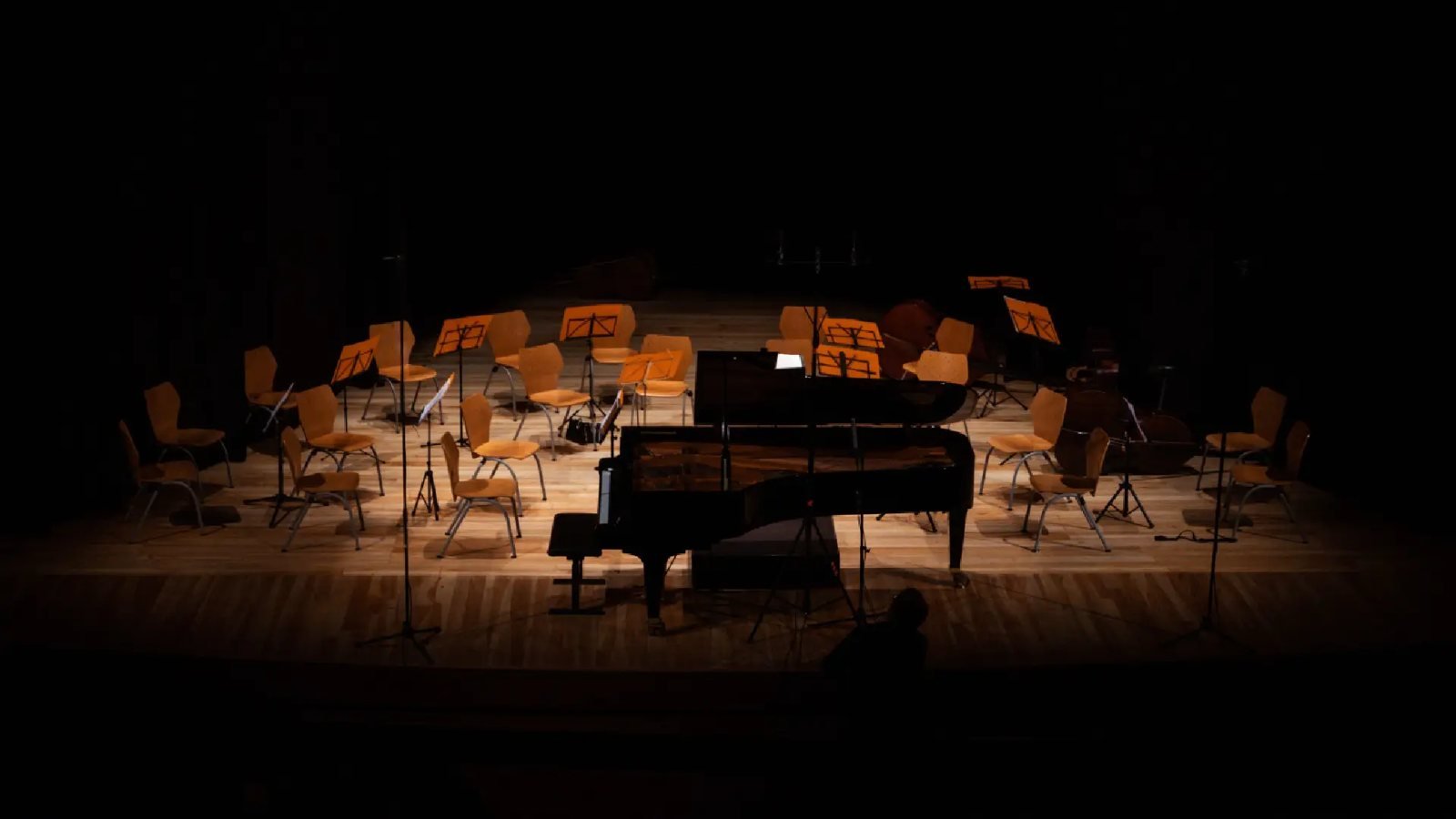 Un piano de cola negro y sillas de madera vacías en un escenario oscuro con atriles iluminados