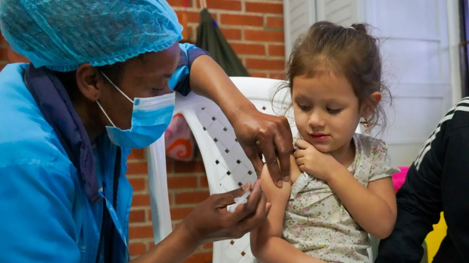 Un personal de salud con mascarilla y gorro quirúrgico vacuna a una niña pequeña en un espacio exterior